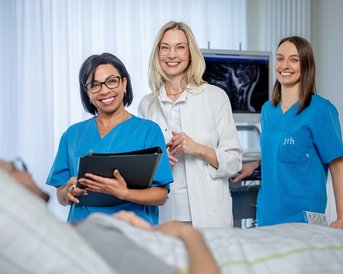 Drei medizinische Fachkräfte in blauen Uniformen stehen lächelnd vor einem Patienten im Krankenhauszimmer, während ein Ultraschallgerät im Hintergrund sichtbar ist.