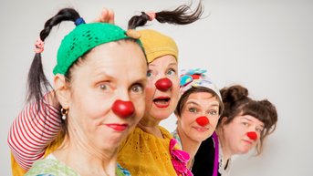 Vier Frauen in bunten Clownskostümen und roten Nasen posieren fröhlich. Sie tragen verschiedene Kopfbedeckungen und haben unterschiedliche Frisuren, während sie in die Kamera schauen.