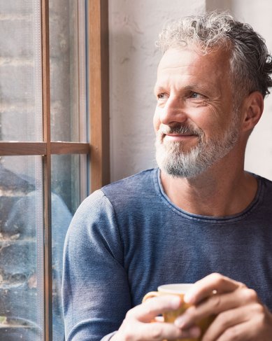 Ein älterer Mann mit grauem Bart und lockigem Haar sitzt am Fenster und lächelt, während er eine Tasse in der Hand hält. Im Hintergrund ist eine helle, gemütliche Innenraumgestaltung zu sehen.
