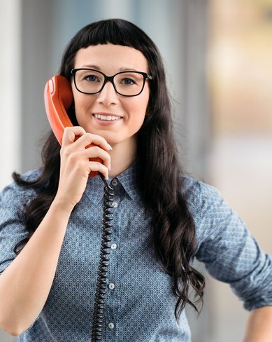 Eine Frau mit langen, dunklen Haaren und Brille lächelt, während sie ein rotes Telefon hält. Sie trägt ein blaues Hemd und steht in einem modernen, hellen Raum mit verschwommenem Hintergrund.
