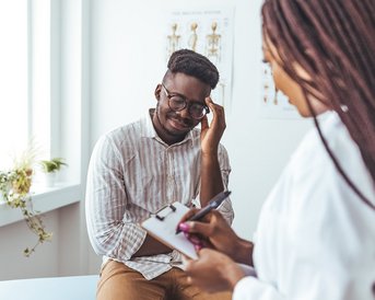 Ein Mann sitzt auf einer Untersuchungsliege und hält sich nachdenklich die Hand an den Kopf. Eine Frau in einem weißen Kittel schreibt Notizen auf einem Clipboard. Anatomische Diagramme sind an der Wand sichtbar.