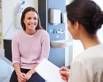 Eine lächelnde Frau sitzt in einem Behandlungsraum und spricht mit einer anderen Frau, die Notizen macht. Die Umgebung ist hell und modern, mit medizinischen Geräten im Hintergrund.