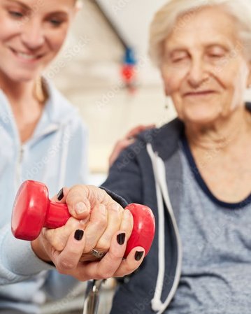 Eine ältere Frau hält ein rotes Hantelgewicht, während eine jüngere Frau sie anleitet. Beide Personen sind in einem Trainingsraum, und die jüngere Frau lächelt, während sie Unterstützung bietet.