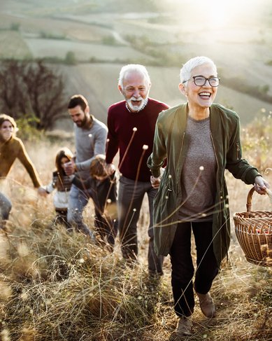 Eine Gruppe von fünf Personen wandert auf einem Hügel, während eine ältere Frau mit einer Korbtasche lächelt. Die Umgebung ist ländlich mit Feldern im Hintergrund und sanften Hügeln.
