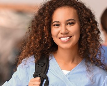 Junge Frau mit lockigem Haar trägt eine hellblaue Uniform und einen Rucksack. Sie lächelt freundlich in die Kamera. Im Hintergrund sind weitere Personen in ähnlicher Kleidung zu sehen.