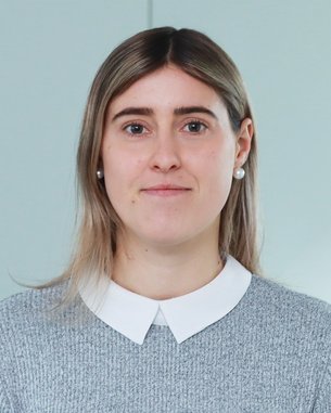 Eine Person mit langen, blonden Haaren und Ohrringen, die in einem grauen Pullover und einem weißen Hemd gekleidet ist, schaut direkt in die Kamera.