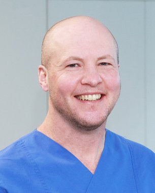 Ein Mann mit Glatze trägt ein blaues medizinisches Outfit und lächelt freundlich in einem hellen Raum.