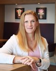 Eine Frau mit langen, blonden Haaren sitzt an einem Tisch in einem modernen Raum, lächelt und trägt ein helles Oberteil.