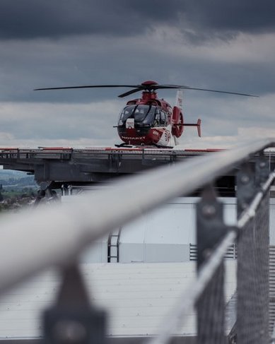Ein Hubschrauber steht auf einer Landeplattform eines Gebäudes, umgeben von bewölktem Himmel und einer ländlichen Landschaft im Hintergrund.