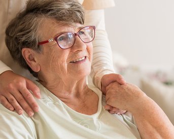 Eine ältere Frau mit Brille sitzt im Rollstuhl, während eine Pflegekraft sie sanft an der Schulter berührt. Im Hintergrund ist ein älterer Mann zu sehen, der mit einem Löffel isst.
