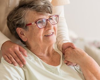 Eine ältere Frau mit Brille sitzt im Rollstuhl, während eine Pflegekraft sie sanft an der Schulter berührt. Im Hintergrund ist ein älterer Mann zu sehen, der mit einem Löffel isst.