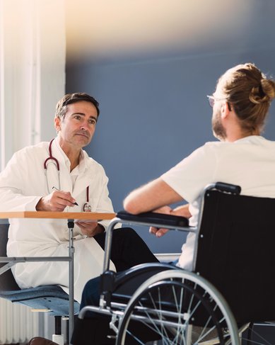 Ein Arzt im weißen Kittel mit Stethoskop sitzt an einem Tisch und spricht mit einem Patienten, der im Rollstuhl sitzt. Beide Personen sind in einem hellen Raum mit Fenstern.