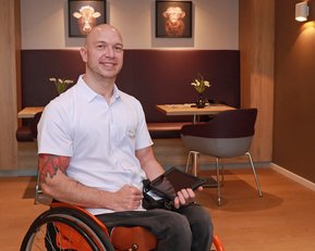 Ein Mann im Rollstuhl sitzt in einem modernen Raum, hält ein Tablet in der Hand und trägt ein weißes Hemd. Im Hintergrund sind zwei Bilder von Kühen und eine Tischanordnung sichtbar.