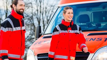 Zwei Rettungssanitäter in roten Uniformen gehen in Richtung eines Rettungswagens. Sie tragen medizinische Ausrüstungen und lächeln. Der Rettungswagen hat orangefarbene und weiße Farben mit der Aufschrift „Rettungsdienst“.