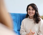 Eine Frau mit braunen Haaren sitzt auf einem Sofa, hält ein Notizbuch und einen Stift, während sie lächelt und aufmerksam zuhört.
