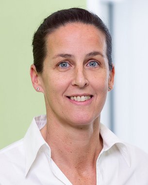 Eine Frau mit kurzen, dunklen Haaren trägt ein weißes Hemd und lächelt. Der Hintergrund ist in einem hellen Grünton gehalten.