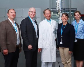 Fünf Personen stehen in einem modernen Raum vor einer transparenten Wirbelsäule. Sie tragen formelle Kleidung und Ausweise. Der zentrale Mann in einem weißen Kittel ist umgeben von drei Männern und einer Frau, die lächeln.