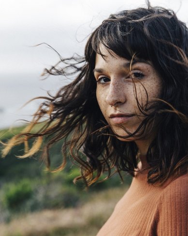 Eine Frau mit lockigem, braunem Haar und Sommersprossen steht im Freien, während der Wind durch ihr Haar weht. Im Hintergrund sind sanfte Hügel und das Meer sichtbar.