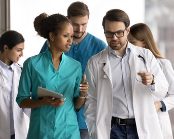 Zwei medizinische Fachkräfte, eine Frau in einem türkisfarbenen Kittel und ein Mann in einem weißen Kittel, diskutieren. Im Hintergrund sind weitere Teammitglieder in medizinischer Kleidung zu sehen, die sich auf den Weg machen.