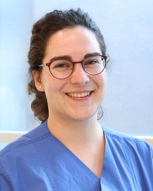Eine Person mit lockigem Haar trägt eine blaue medizinische Uniform und Brille. Sie lächelt freundlich und steht vor einem Fenster mit natürlichem Licht.