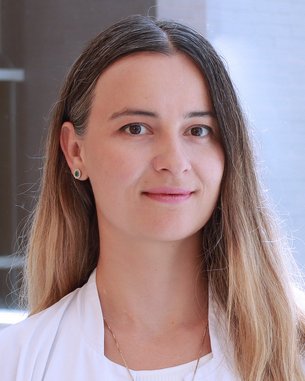 Eine Person mit langen, braunen Haaren trägt einen weißen Laborkittel und steht in einem modernen Innenraum. Sie hat einen neutralen Gesichtsausdruck und trägt eine Halskette.