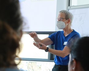 Eine Person mit Brille und Maske erklärt etwas vor einer Gruppe von Zuhörern in einem modernen Raum. Ein Flipchart ist im Hintergrund sichtbar.