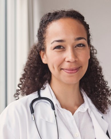 Eine Ärztin mit lockigem Haar trägt einen weißen Kittel und ein Stethoskop um den Hals. Sie steht in einem hellen Raum und lächelt freundlich.