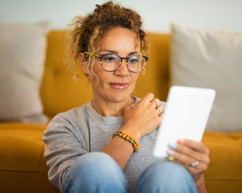 Eine Frau mit lockigem Haar und Brille sitzt auf einem Sofa und betrachtet ein Tablet. Sie trägt ein graues Sweatshirt und eine bunte Armbanduhr. Der Hintergrund zeigt ein gelbes Sofa mit Kissen.