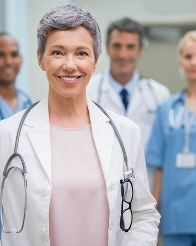 Eine lächelnde Ärztin mit grauen Haaren trägt einen weißen Kittel und ein Stethoskop. Im Hintergrund stehen drei weitere medizinische Fachkräfte in blauer Kleidung. Die Szene spielt in einer Klinik oder Arztpraxis.