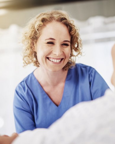 Eine Pflegekraft mit lockigem, blondem Haar lächelt freundlich zu einem älteren Patienten, der im Bett liegt. Die Szene vermittelt eine Atmosphäre der Fürsorge und Unterstützung im Gesundheitswesen.