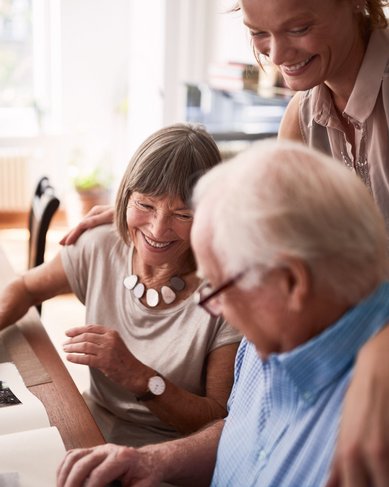 Eine ältere Frau und ein älterer Mann blättern gemeinsam durch ein Fotoalbum, während eine jüngere Frau neben ihnen steht und lächelt. Der Tisch ist mit einer Vase und weiteren Gegenständen dekoriert.