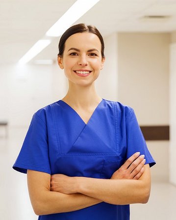 Eine Frau in blauer medizinischer Kleidung steht mit verschränkten Armen in einem hellen Krankenhausflur. Sie lächelt freundlich und wirkt professionell. Der Flur ist schlicht und gut beleuchtet.