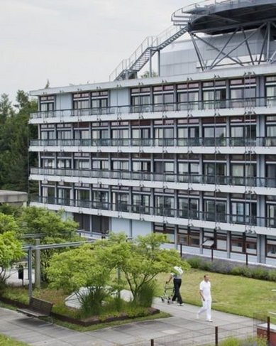 Ein modernes Gebäude mit mehreren Etagen, umgeben von begrünten Terrassen und Bäumen. Auf einer der Terrassen sind zwei Personen zu sehen, die sich in einem Gartenbereich aufhalten.