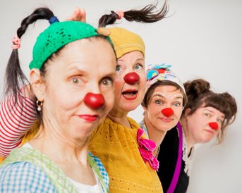 Vier Frauen in bunten Clownskostümen und roten Nasen posieren fröhlich. Sie tragen verschiedene Kopfbedeckungen und haben unterschiedliche Frisuren, während sie in die Kamera schauen.