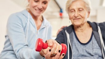 Eine ältere Frau hält ein rotes Hantelgewicht, während eine jüngere Person ihr assistiert. Beide sind in einem Trainingsraum, und die jüngere Person zeigt eine unterstützende Haltung.