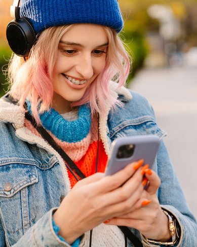 Eine junge Frau mit Kopfhörern und einer blauen Mütze schaut auf ihr Smartphone. Sie trägt eine Jeansjacke und einen bunten Pullover. Die Umgebung zeigt eine städtische Straße mit parkenden Autos im Hintergrund.
