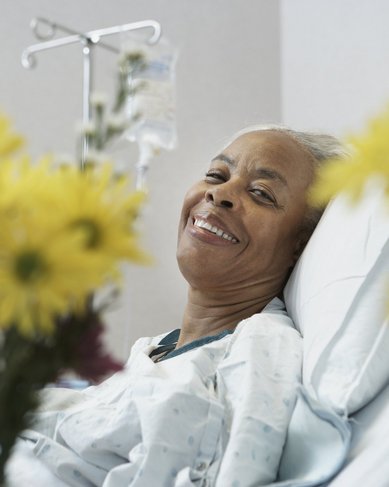 Eine lächelnde ältere Frau liegt in einem Krankenhausbett, umgeben von bunten Blumen. Sie trägt ein Krankenhaushemd und wirkt entspannt. Ein Infusionsständer ist im Hintergrund sichtbar.