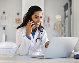 Eine Ärztin spricht am Telefon, während sie auf einen Laptop schaut. Sie trägt einen weißen Kittel und hat ein Stethoskop um den Hals. Im Hintergrund ist ein medizinisches Modell sichtbar.