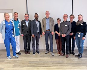 Eine Gruppe von zehn Personen steht in einem Raum, einige tragen Arbeitskleidung. Sie lächeln und posieren für ein Foto, während sie sich in einer professionellen Umgebung befinden. Namensschilder sind sichtbar.