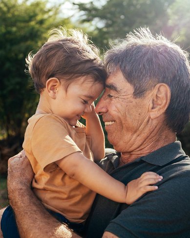 Ein älterer Mann hält ein Kleinkind in den Armen, während sie sich anlächeln und ihre Stirnen berühren. Die Szene spielt sich in einer natürlichen Umgebung mit Bäumen im Hintergrund ab.