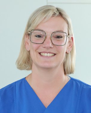 Eine Person mit blonden Haaren und großen Brillenläsern trägt einen blauen Arztkittel und lächelt freundlich in die Kamera. Der Hintergrund ist hell und neutral.